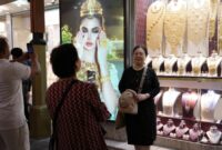 Para turis berpose untuk foto di depan toko perhiasan dengan layar tampilan yang menampilkan gambar model yang mengenakan perhiasan emas yang memukau.