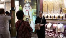 Para turis berpose untuk foto di depan toko perhiasan dengan layar tampilan yang menampilkan gambar model yang mengenakan perhiasan emas yang memukau.