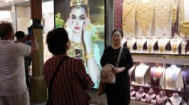 Para turis berpose untuk foto di depan toko perhiasan dengan layar tampilan yang menampilkan gambar model yang mengenakan perhiasan emas yang memukau.